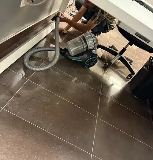 A person vacuuming the floor underneath a white office desk.