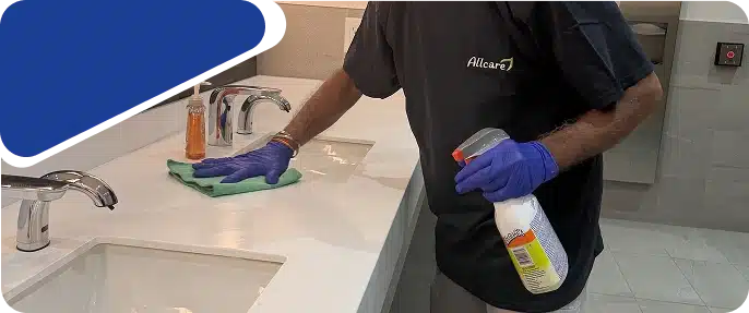 A cleaner wearing blue gloves wiping a surface with a green cloth and spray bottle.
