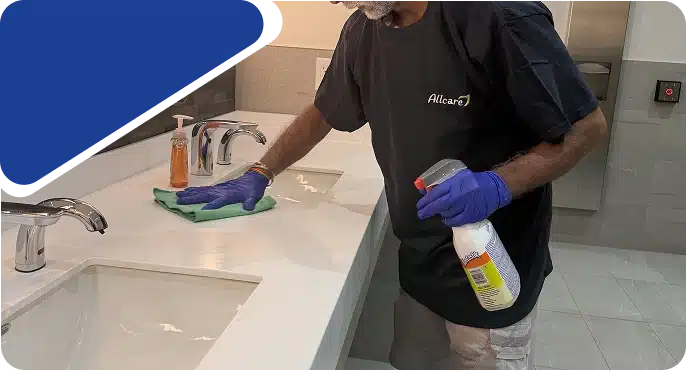 A person wearing blue gloves wiping a white counter with a green cloth and a spray bottle.